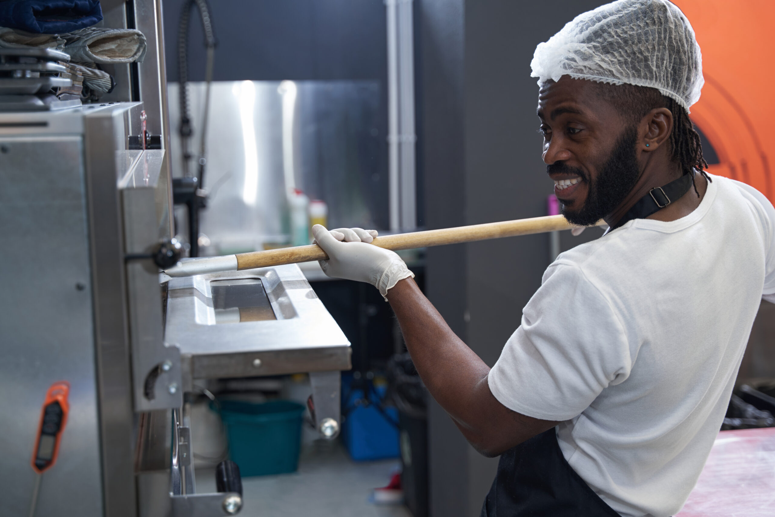 Chef in uniform and protective gloves standing in the kitchen, opens the oven door and putting pastries inside